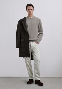 Szary sweter, ciemny płaszcz z dużymi guzikami, jasne dopasowane spodnie i ciemne botki za kostkę, na neutralnym tle.