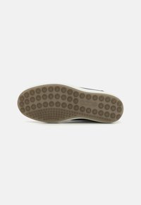 Semelle d'une chaussure de sport, présentant un caoutchouc lisse de couleur marron clair avec des motifs de grip circulaires, contrastant avec une intercalaire blanche.