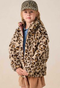 Meisje in een luipaardprint cap en bijpassende fluffy luipaardprint jas, met een blauwe shirt en een beige corduroy rok.