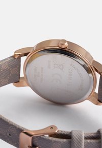 Klocka i roséguld av rostfritt stål med ett mönstrat textilarmband. Har ett runt boett, krona och en genomskinlig baksida som visar varumärkesdetaljer.