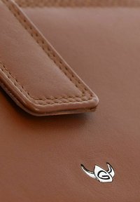 Gros plan du coin d’un sac en cuir marron avec une couture détaillée et un petit logo argenté ressemblant à un emblème stylisé à cornes.