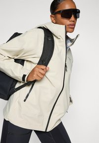 Personne portant une veste beige clair et de grandes lunettes de soleil noires, portant un sac à dos noir, posant devant un fond uni.