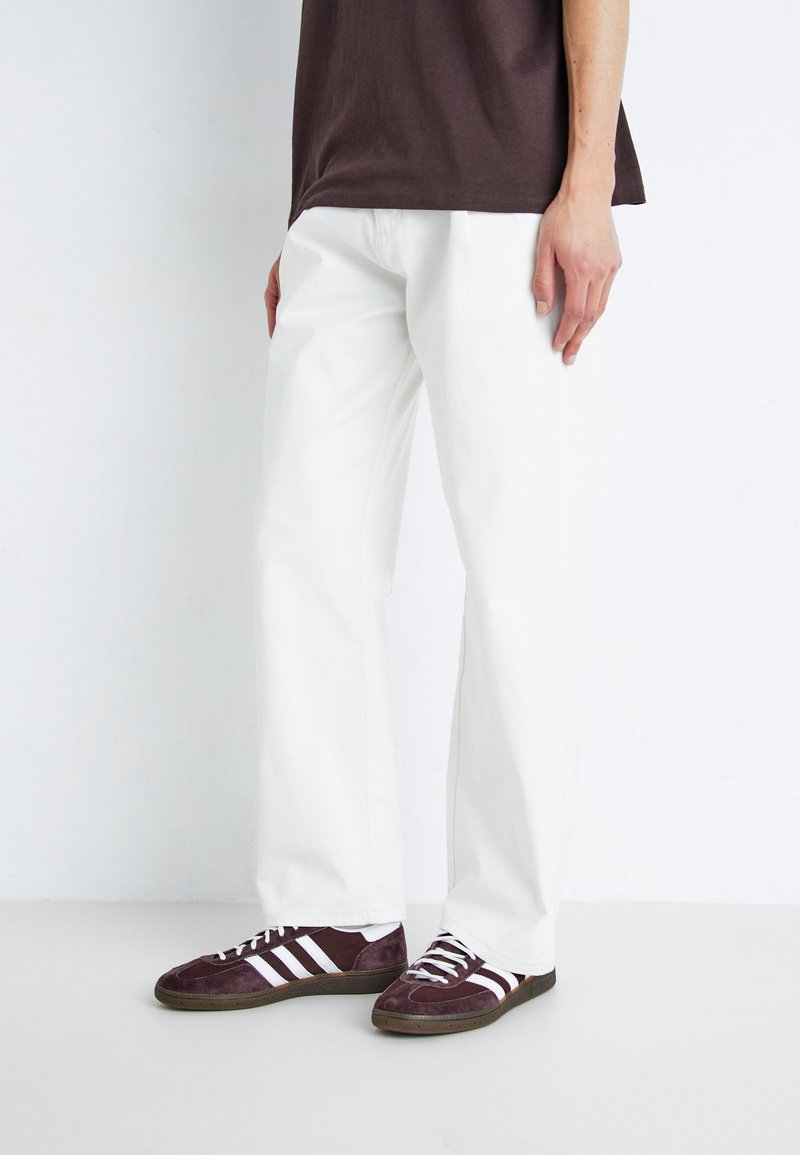 Witte katoenen broek met een rechte pijp, gecombineerd met bordeauxrode schoenen met witte strepen. De outfit wordt compleet gemaakt met een bruine t-shirt.