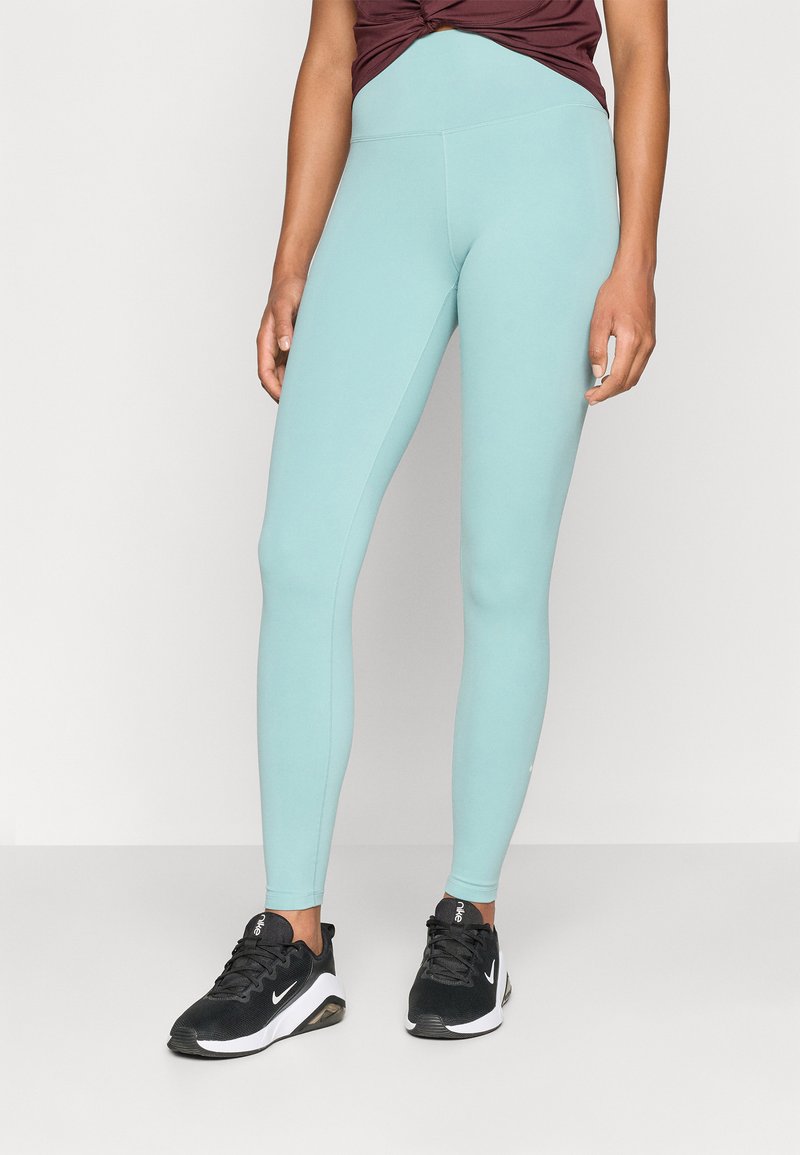 Helle türkisfarbene Leggings mit glatter Textur und hohem Taillenbund, kombiniert mit schwarzen Sportschuhen mit weißen Akzenten.