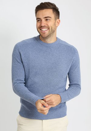 Man lacht, draagt een lichtblauwe gebreide trui en beige broek, terwijl hij de mouwmanchetten aanpast tegen een eenvoudige witte achtergrond.