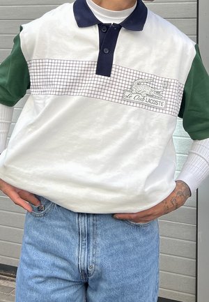 White and green short-sleeve polo with navy collar, grid pattern, and embroidered crocodile logo labeled "Le Club Lacoste." Worn with light jeans.