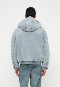 Hombre de pie de espaldas, luciendo una chaqueta con capucha de denim azul claro y jeans desgastados de color azul, frente a un fondo blanco liso.
