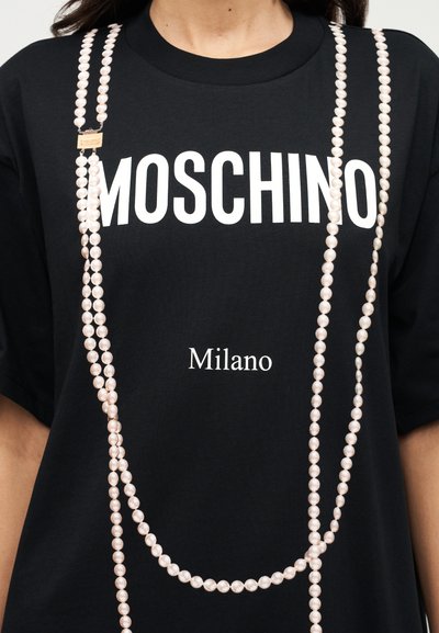 Μαύρο T-shirt με το λευκό τυπώμα "MOSCHINO" και "Milano"; διαθέτει αλυσίδες που μοιάζουν με μαργαριτάρια κρεμασμένες μπροστά για παραπάνω λεπτομέρεια.