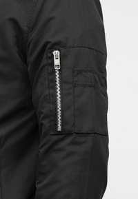 Gros plan sur la manche noire d'un pantalon avec une fermeture éclair verticale argentée et deux poches cousues sur le bras, sur un fond clair.