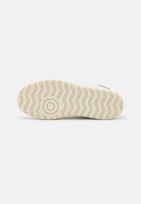 Semelle d'une chaussure New Balance beige clair avec des motifs ondulés et un logo circulaire près du talon sur fond blanc.