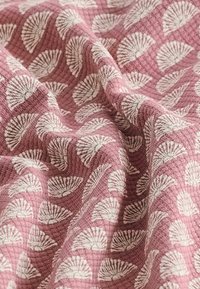 Tissu avec un fond texturé rose ornée de motifs de coquillages blancs, créant un design subtil et répétitif. Tissu doux et tissé.