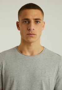 Szürke rövid ujjú póló, kerek nyakkivágással, puha textúrával, a bal mellrészen egy kis hímzett négyzettel.