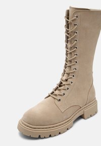 Beige suede knee-high lace-up boots with a thick, ridged sole and metal eyelets, featuring a smooth texture and sturdy construction.