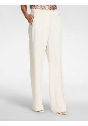 Donna che indossa pantaloni crema a gamba larga, in piedi con le mani nelle tasche, abbinati a un top di paillettes e tacchi aperti.