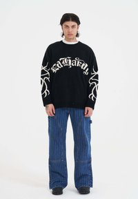 Młoda osoba z średniej długości ciemnymi włosami, nosząca czarny sweter z białymi wzorami i szerokie niebieskie spodnie w prążki na białym tle.