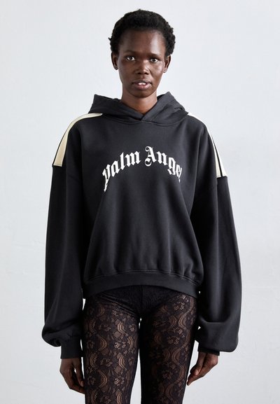 Hoodie noir surdimensionné avec le logo "Palm Angels" en blanc, doté d'accents crème sur les épaules. Associé à des leggings en dentelle noirs.