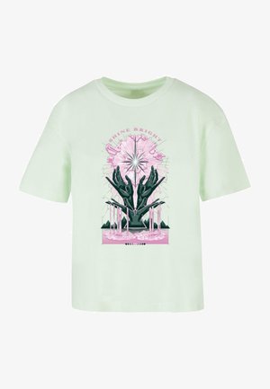 Jasnozielona bawełniana koszulka z graficznym wzorem, przedstawiającym czarne dłonie, różowe akcenty oraz tekst "ŚWIEC W POSZUKIWANIU" nad promieniującą gwiazdą.