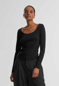 Musta pitkähihaisuus paita, jossa on leveä pyöreä kaula-aukko, valmistettu sileästä ja joustavasta kankaasta. Yhdistetty musta-valkoisiin raidallisiin housuihin.