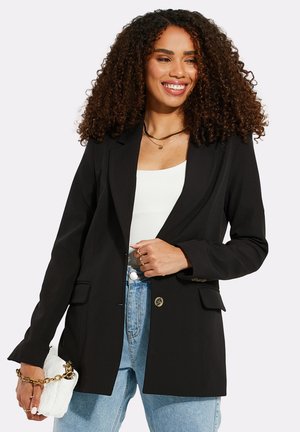 Černý blazer s přiléhavým střihem, dvěma předními kapsami, klopovým límcem, zapínáním na knoflík a hladkým materiálem, nošený přes bílé tričko a světle modré džíny.