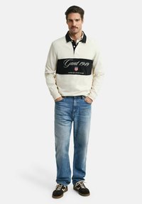 Polo à manches longues couleur crème avec une rayure horizontale noire arborant le logo "Gant 1949", assorti à un jean bleu clair et des baskets foncées.