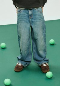 Vide, lyseblå denimjeans med falmede områder og en lett tekstur. Brune joggesko med hvite detaljer. Grønne tennisballer på en grønn overflate.