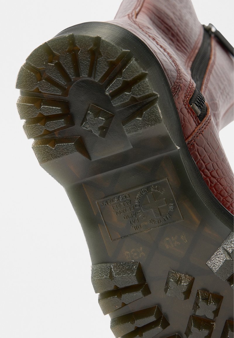 Red textured boot sole with deep traction pattern and angular indentations. Features embossed logo and rugged outsole for grip.