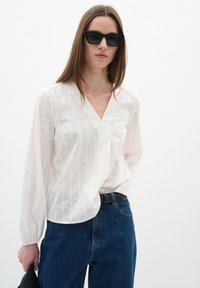 Blusa bianca con scollo a V, righe testurizzate e maniche lunghe. Abbinata a jeans blu a vita alta e una cintura nera, con una borsa nera.