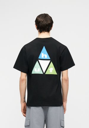 Άτομο που φοράει μαύρο μπλουζάκι με σχέδιο από τρίγωνα σε μπλε, λευκό και πράσινο χρώμα και γράμματα "h," "u," και "f" στην πλάτη, συνδυασμένο με ανοιχτό γκρι σορτς.