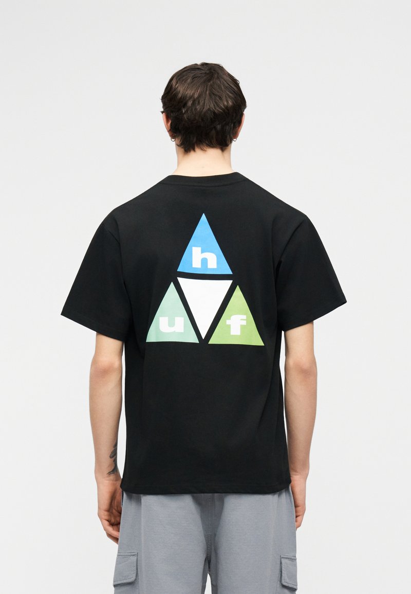 Άτομο που φοράει μαύρο μπλουζάκι με σχέδιο από τρίγωνα σε μπλε, λευκό και πράσινο χρώμα και γράμματα "h," "u," και "f" στην πλάτη, συνδυασμένο με ανοιχτό γκρι σορτς.