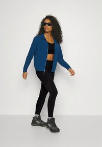 Giacca blu con zip realizzata in materiale morbido, top da allenamento nero, leggings neri con dettagli testurizzati e scarpe da ginnastica nere. Capelli ricci e occhiali da sole.