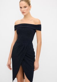 Abito nero scollato sulle spalle in un tessuto testurizzato, con un corpetto aderente, gonna avvolgente con uno spacco alto e una sottile lucentezza.
