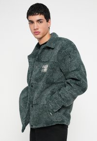 Πράσινο μπουφάν με χνουδωτή επιφάνεια, μαύρα κουμπιά και ένα μικρό λογότυπο. Διαθέτει γιακά και πλαϊνές τσέπες.