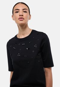 Haut noir à manches courtes avec des appliqués floraux et des sequins. Tissu doux avec un col arrondi et des détails décoratifs sur le devant.