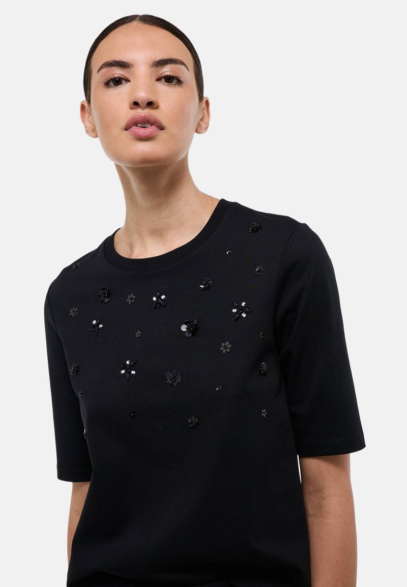 Haut noir à manches courtes avec des appliqués floraux et des sequins. Tissu doux avec un col arrondi et des détails décoratifs sur le devant.