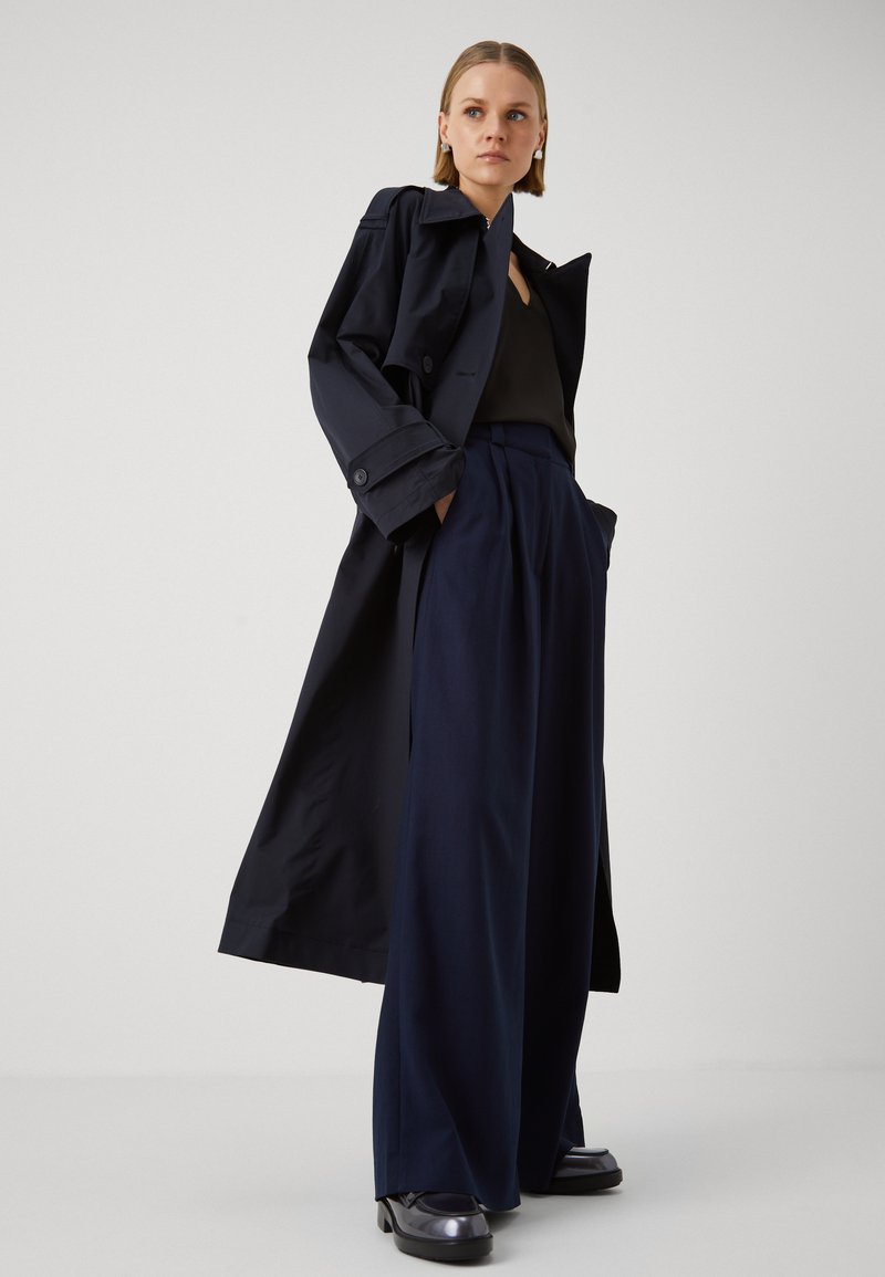 Trench coat blu scuro in un tessuto resistente all'acqua, abbinato a pantaloni blu scuro dalla vestibilità ampia. Texture liscia con chiusura a bottone e gambe larghe.