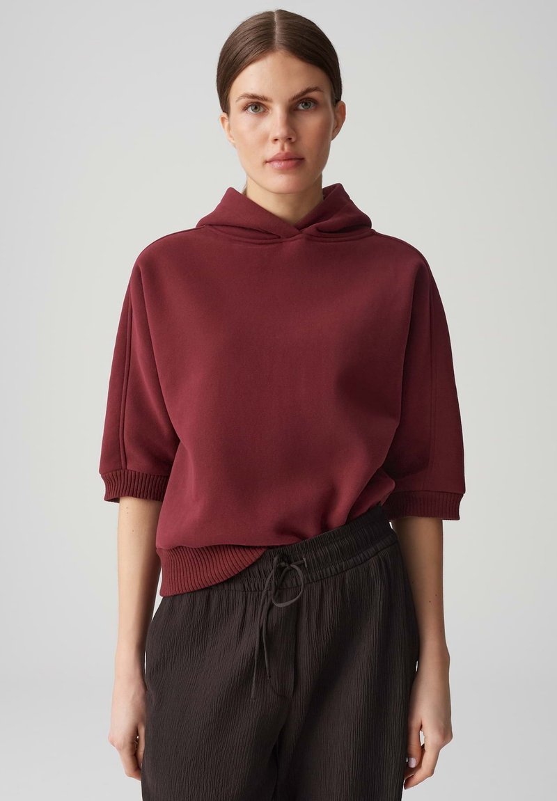 Felpa con cappuccio color bordeaux a maniche corte, con polsini e orlo a coste. Abbinata a pantaloni neri texturizzati con vita elasticizzata.