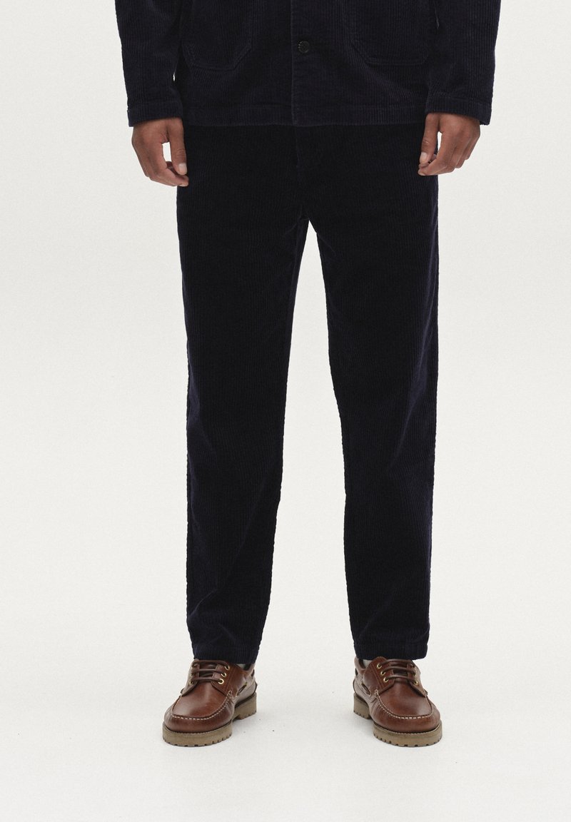 Marineblauwe corduroy jas met knoopsluiting, voorzakken, gecombineerd met bijpassende corduroy broek en bruine leren schoenen met een gestructureerde zool.