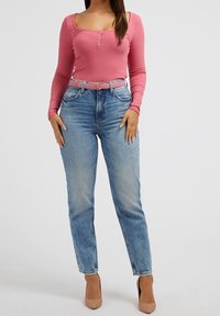 Donna che indossa un top rosa a maniche lunghe, jeans blu a vita alta con una cintura rosa e scarpe con tacco beige, in piedi su uno sfondo bianco.
