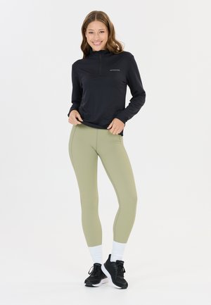 Kvinde smilende, stående med hænderne på hofterne, iført sort langærmet top, lysegrønne leggings, hvide sokker og sorte løbesko.