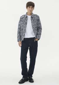 Mladenič oblečen v vzorčasto sivo jakno, belo majico, temno modre kavbojke in črne čevlje z resicami, stoji z eno roko v žepu.