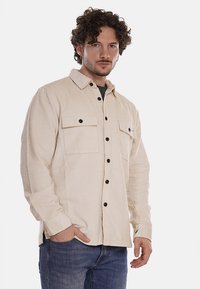 Camicia in corduroy beige con due tasche sul petto, bottoni neri e colletto a punta, abbinata a jeans blu. La texture è rigata e morbida.