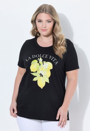 Femme aux cheveux blonds ondulés portant des boucles d'oreilles créoles dorées et un t-shirt noir avec des citrons jaunes et le texte « La Dolce Vita », associée à un pantalon blanc.