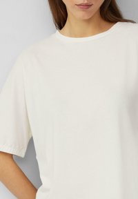 Krótki rękaw, kremowa koszulka wykonana z miękkiej bawełny. Posiada okrągły dekolt i luźny krój z podwiniętymi mankietami.