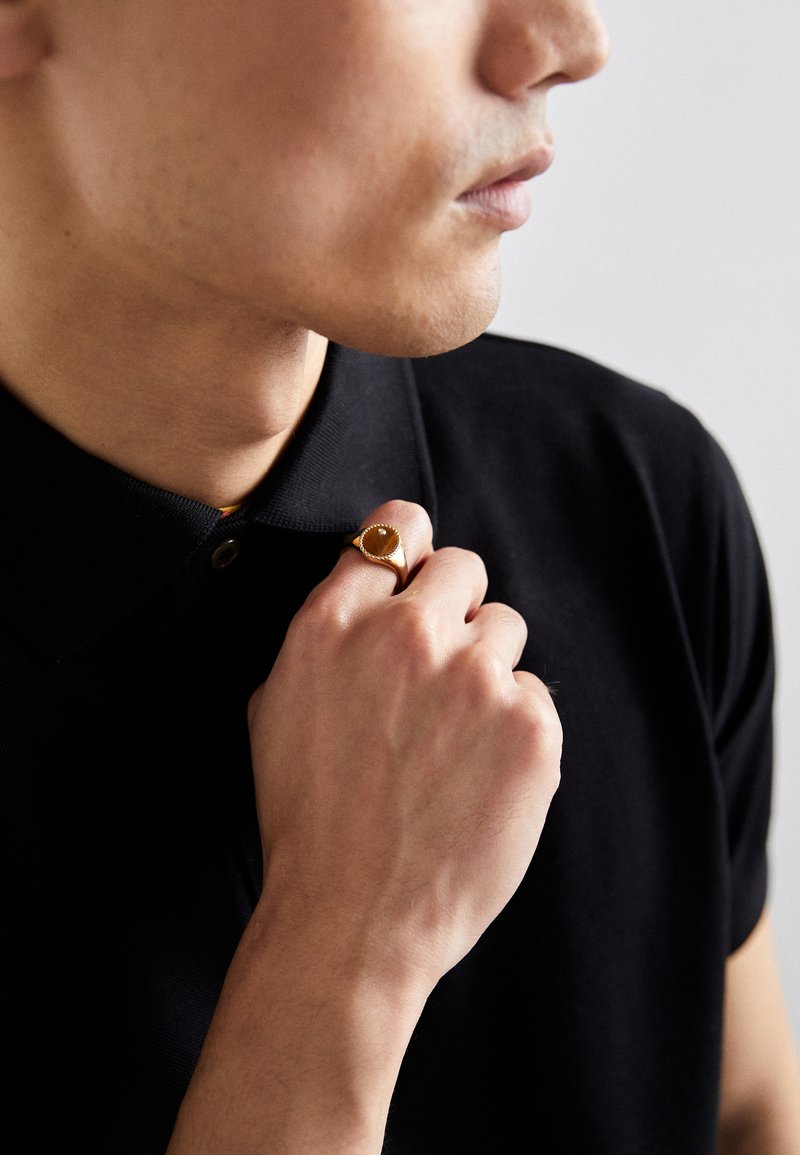 Gold ring with oval amber stone, worn on the right hand. The wearer has a black polo shirt, showing a textured collar and button.