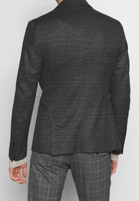 Schwarzes, strukturiertes Blazer mit taillierter Passform, ausgestattet mit einem Schlitz hinten und geknöpften Manschetten, kombiniert mit grauen karierten Hosen.