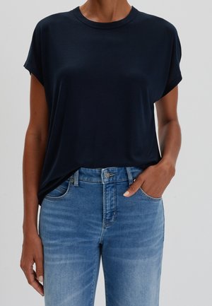 Osoba ubrana w gładki czarny t-shirt z krótkim rękawem i niebieskie dżinsy stoi z jedną ręką w kieszeni na neutralnym tle.