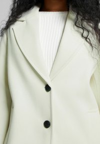 Lätt, krämfärgad blazer med en mjuk textur, med kavajslag och två svarta knappar, lager på en vit ribbad tröja.