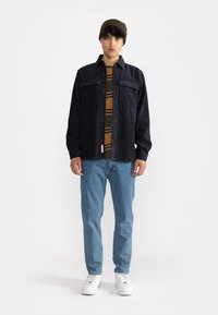Mørkeblå kordfløjl skjortejakke med to brystlommer over en brun og grøn stribet skjorte. Lyseblå jeans og hvide sneakers fuldender looket.
