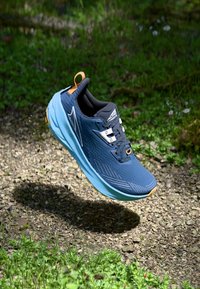 Blauer Laufschuh mit strukturiertem Obermaterial, schwarzer Innenauskleidung und orangefarbenem Akzent. Mit weißen Schnürsenkeln und Logo an der Seite, vor einem natürlichen Hintergrund.