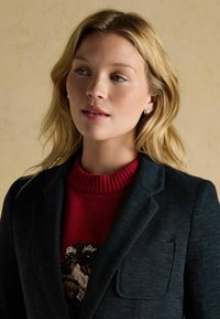 Tmavo zelený herringbone blejzer prehodený cez červený sveter so vzorom psíkov. Obsahuje zárezový golier a náprsní vrecko. Hladká textúra, priliehavý dizajn.